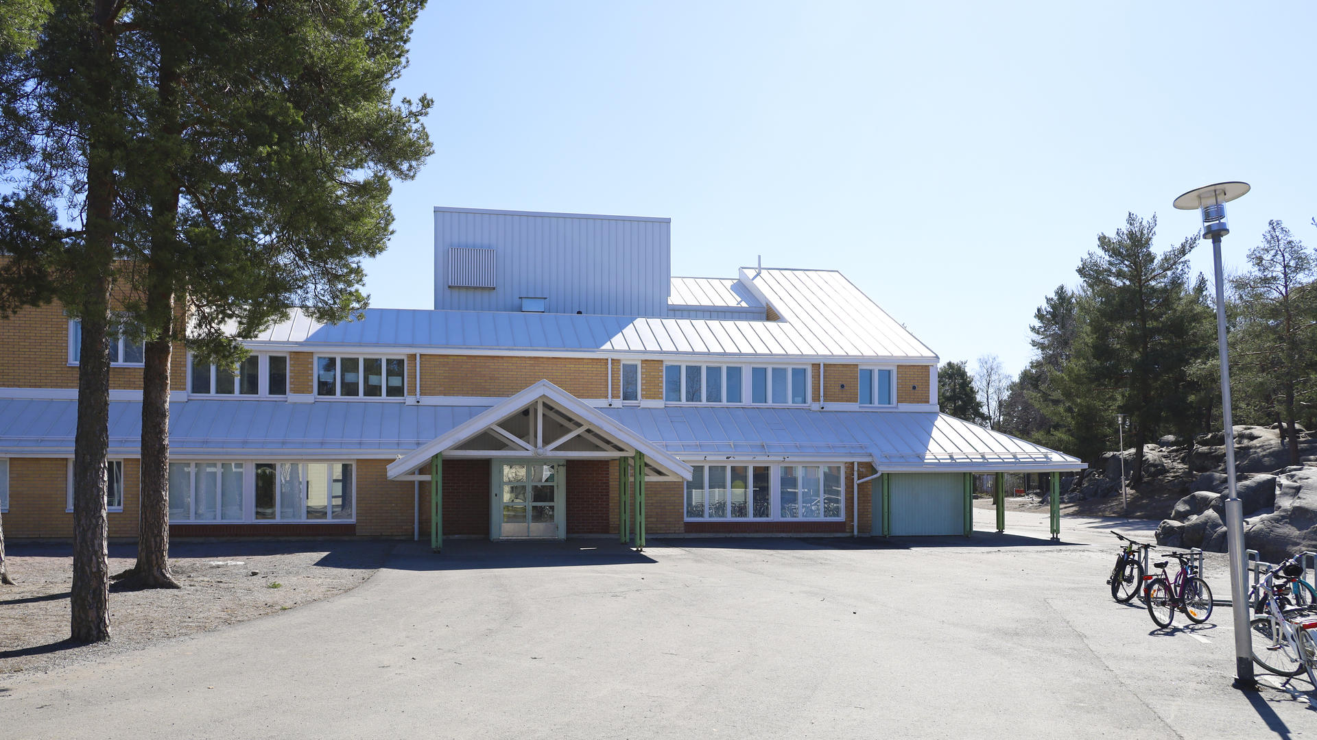 Friisilän koulun piha. Keltaisen matalan rakennuksen edessä asfalttipiha ja puita.