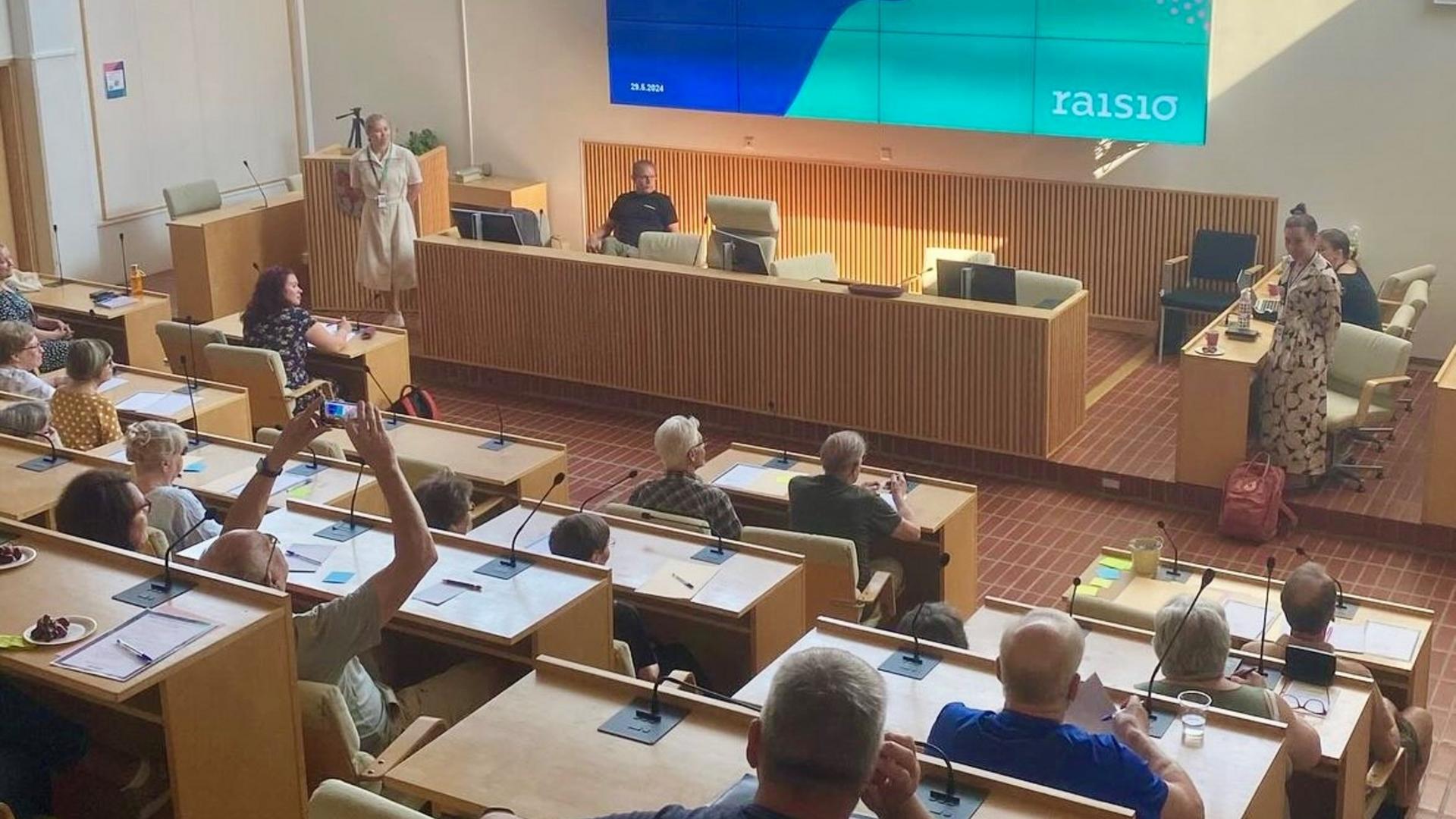 Joukko ihmisiä valtuustosalissa istumassa. 