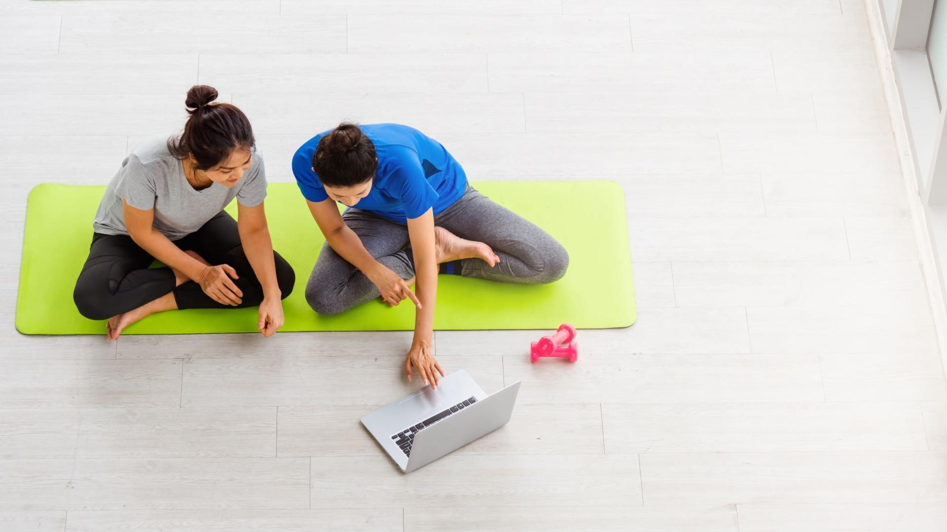 Kaksi naista istumassa vihreällä joogamatolla katsomassa kannettavan tietokoneen ruutua.