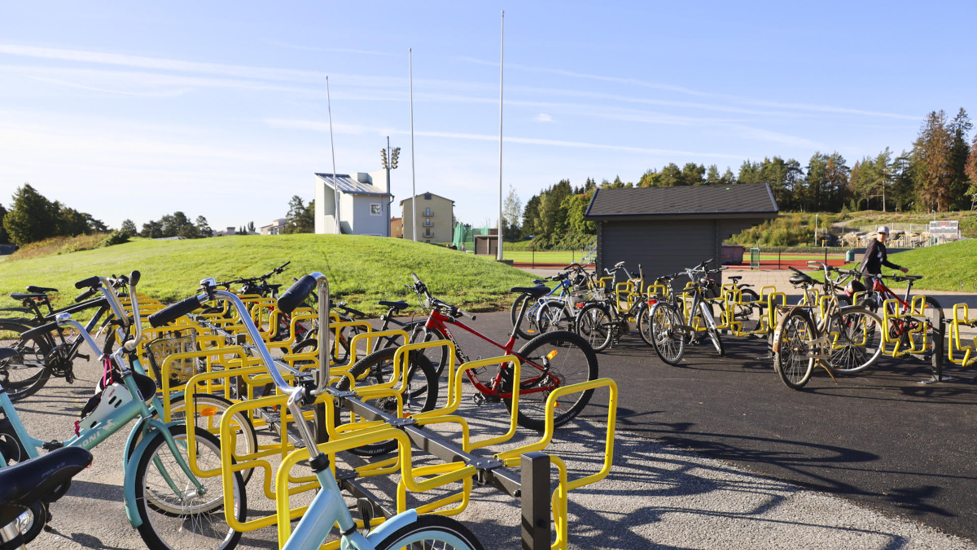 Keltaisia runkolukittavia pyörätelineitä Kerttulan liikuntakeskuksen pihassa. Taustalla Kerttulan yleisurheilukenttä. 