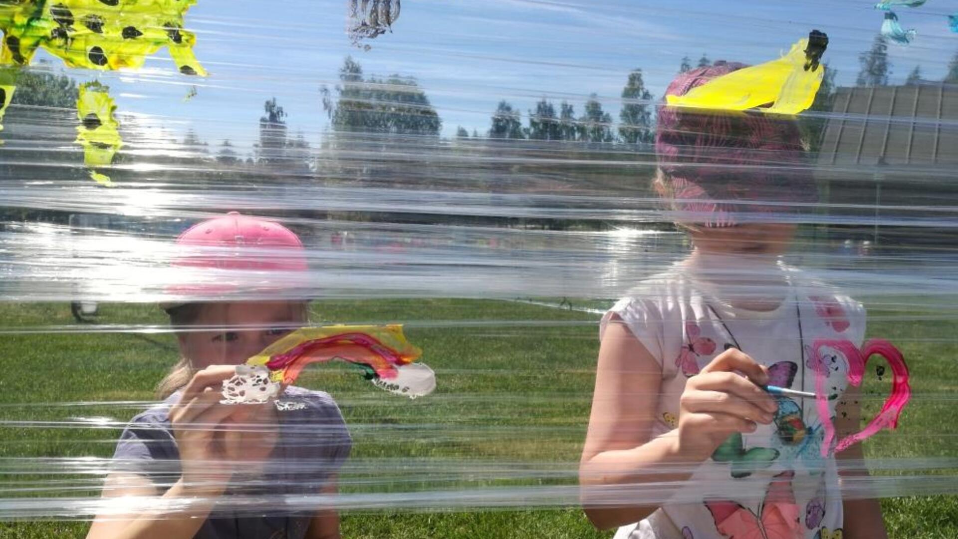 Lapset maalaavat läpinäkyvää muovia aurinkoisessa kesäsäässä.