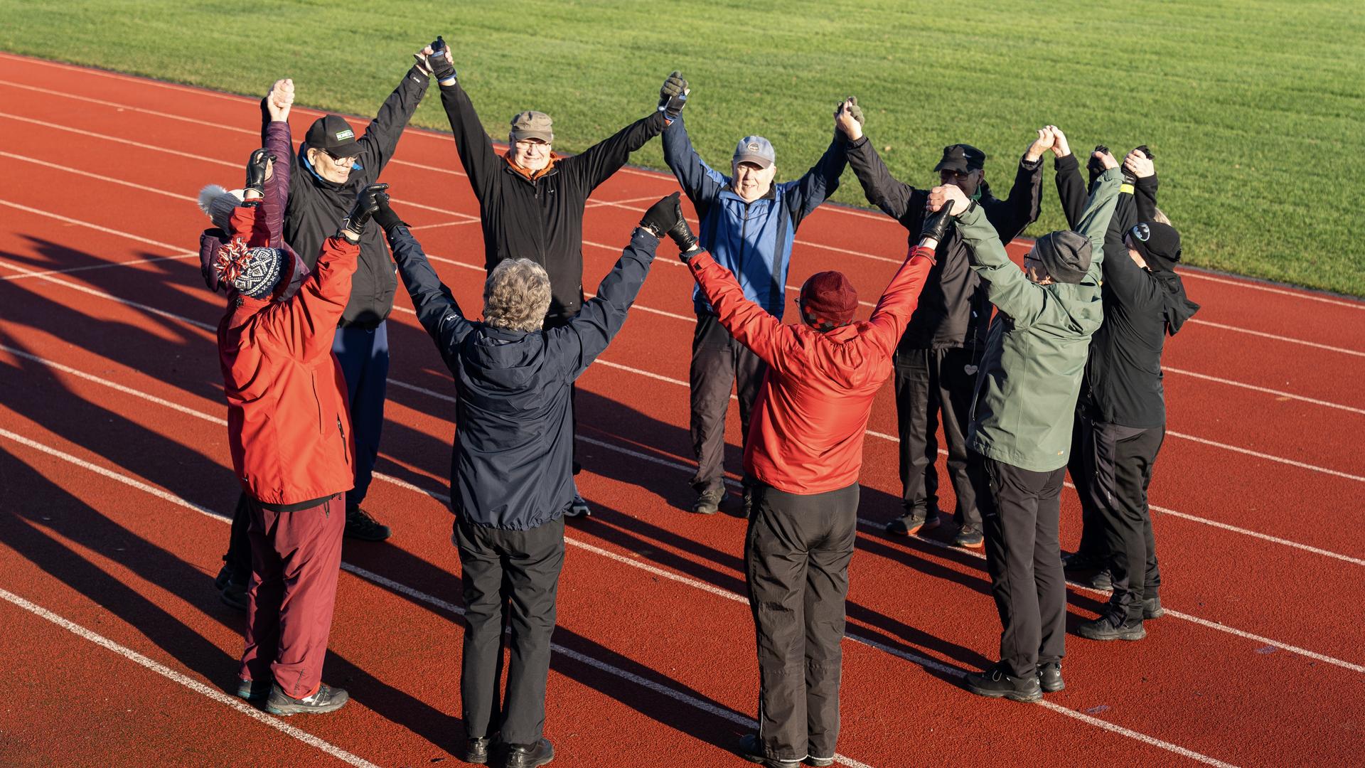 Joukko eläkeläisiä seisomassa yleisurheilukentällä piirissä kädet yhdessä ylhäällä. 
