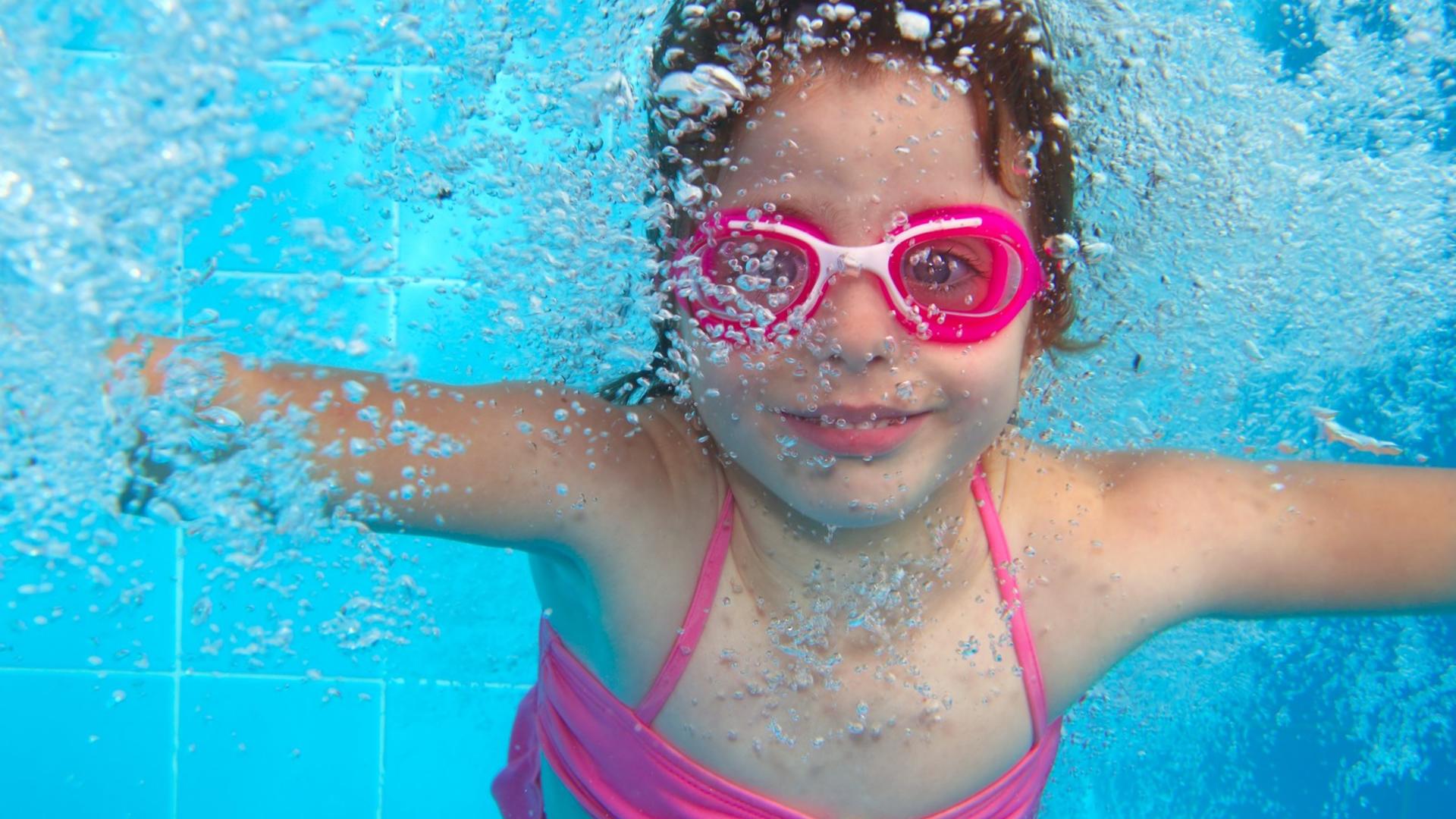 Vaalenapunaiseen uimapukuun ja uimalaseihin pukeutunut tyttö sukeltaa uima-altaassa.