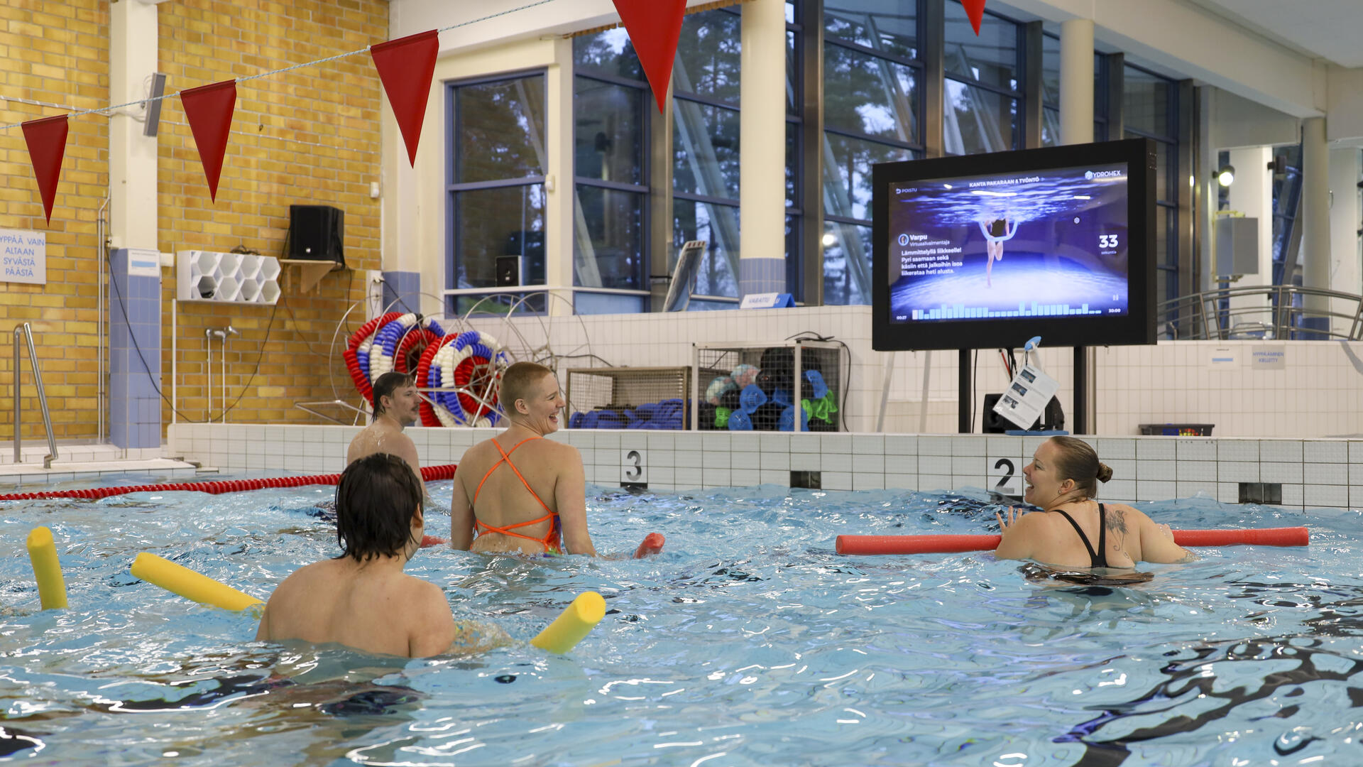 joukko iloisia ihmisiä tekemässä virtuaalista vesijumppaa uima-altaassa.