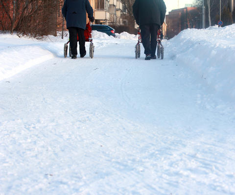 Kaksi henkilöä kulkee rollaattorilla lumisella kadulla.