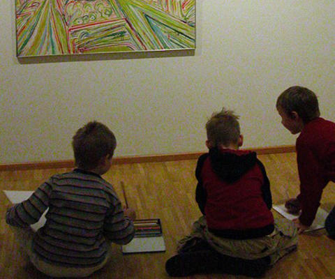 Kolme lasta istuu lattialla taideteoksen edessä näyttelyssä.