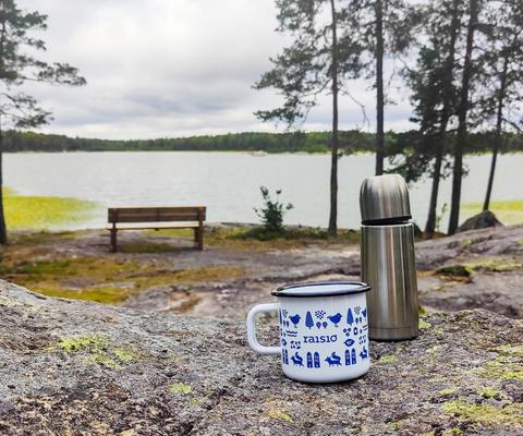 Penkki ja puita Haunisten altaan rannalla, kuvan etualalla Raisio-aiheinen peltimuki ja termospullo kalliolla. 