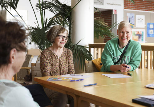 Vanhempi nainen kukikkaassa paidassa ja nuorempi vaaleatukkainen nainen vihreässä paidassa hymyilevät pöydän äärellä istuen.