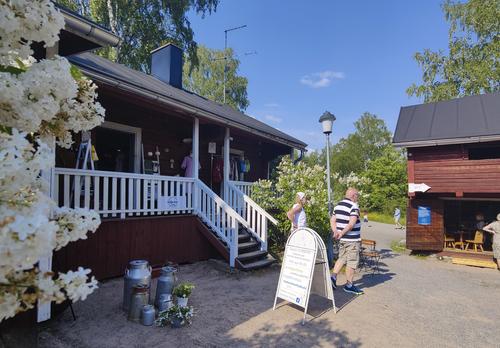 Krookilan kotiseutukeskuksen pihapiiri, jossa punaiset puurakennukset. Rakennuksen edessä kyltti ja kaksi ihmistä sekä kukkiva puu tai pensas.