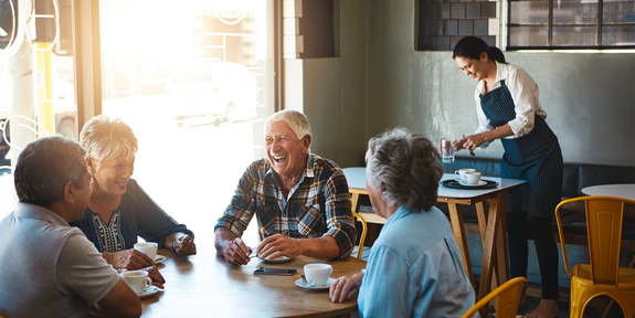 Neljä seniori-ikäistä henkilöä kahvilassa rupattelemassa. Taustalla kahvilatyöntekijä.