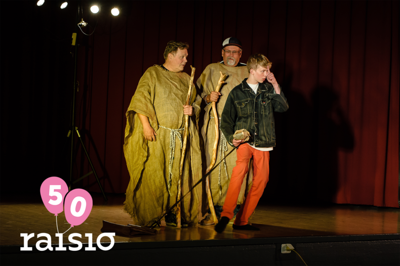 Lavalla kolme näyttelijää. Vasemmassa alareunassa Raisio-logo ja kaksi pinkkiä ilmapalloa.