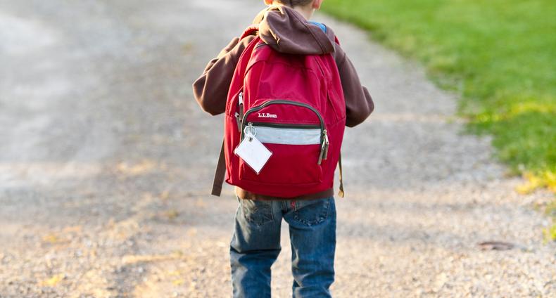 Pieni poika kävelee hiekkatiellä reppu selässä.