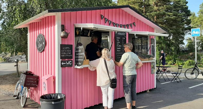 Vaaleanpunainen pieni kahvilakoju, jonka edessä kaksi henkilöä selkä kameraan päin