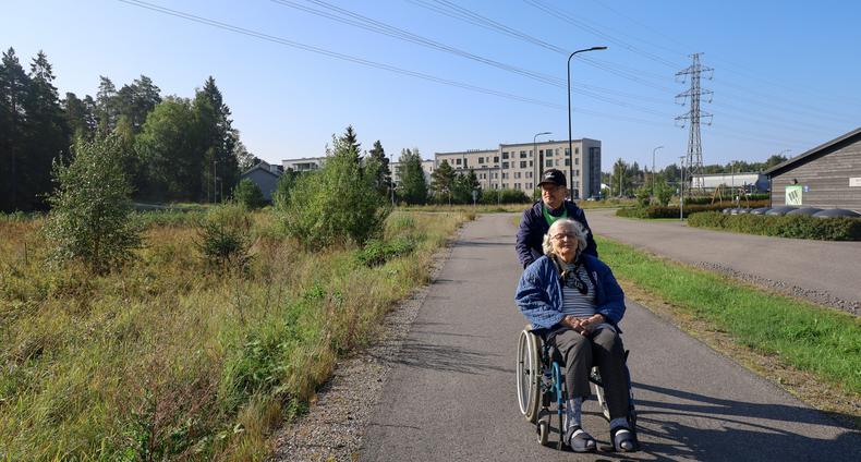 Vapaaehtoinen ulkoiluystävä työntää ikäihmistä ulkona pyörätuolissa.